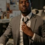Businessman adjusting tie at office desk with documents, conveying confidence and professionalism.