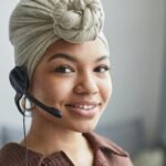 Delighted African American female call agent with headset wearing turban looking away while standing in light room on blurred background