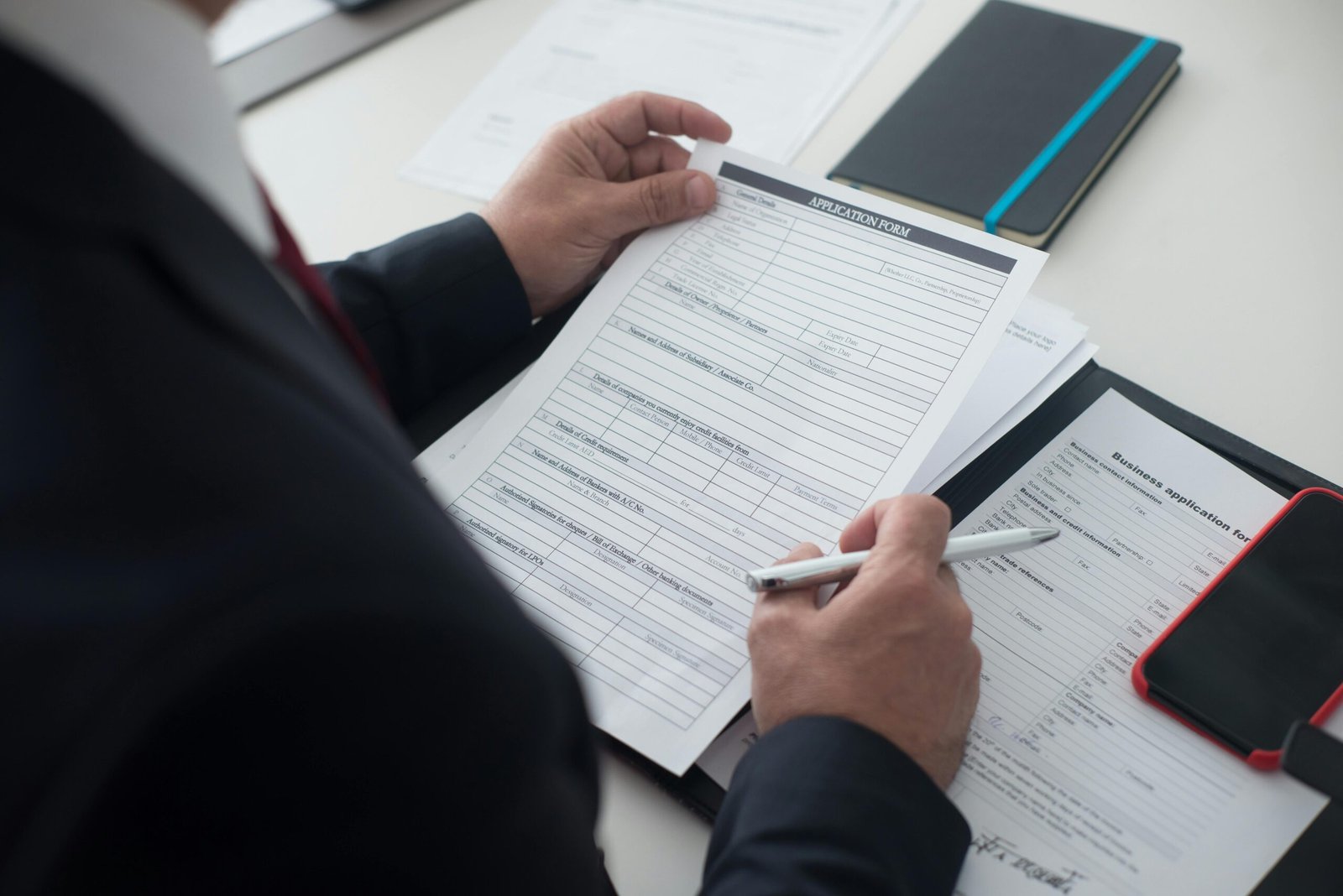 A businessman in a black suit reviews a printed application form with a pen on his desk.
