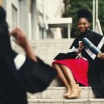 Black female graduate in gown smiles with diploma on city stairs, embodying achievement.