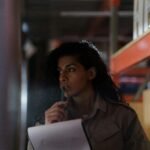 A woman in a warehouse contemplatively checking inventory under low light conditions.