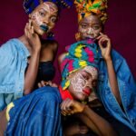 Three women in colorful African outfits with face paint and headdresses celebrating culture.